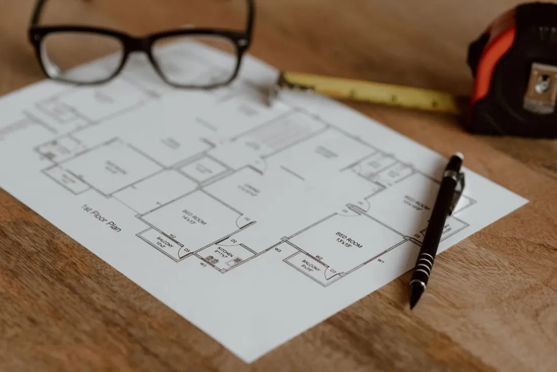 A printed house floor plan on a desk with a pen, glasses and tape measure